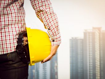 Hand's engineer worker holding yellow safety helmet with building on site background.