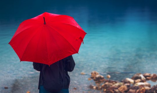 Woman with red umbrella contemplates on rain in front of a lake. Sad and lonely female person looking into distance.